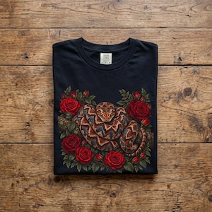 Può includere: Una t-shirt nera con una stampa grafica di un serpente marrone e beige avvolto su un ramo, circondato da vivaci rose rosse e foglie verdi. L'etichetta interna sul collo riporta il testo Explore Course e la taglia L.
