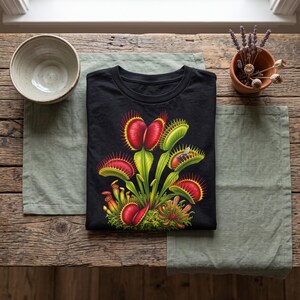 Puede incluir: Una camiseta negra con un estampado gráfico vibrante de varias plantas atrapamoscas con hojas de color rojo brillante y verde, bordes espinosos y un pequeño insecto capturado dentro de una de las trampas sobre la tela oscura.