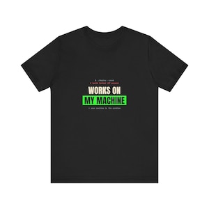 Könnte beinhalten: Ein schwarzes Kurzarm-T-Shirt mit dem Text "WORKS ON MY MACHINE" in Weiß und Grün, ergänzt durch kleinen Text im Terminal-Stil in Weiß und Rot ober- und unterhalb, einschließlich "$ ./deploy --prod" sowie "X build failed" als Fehlermeldung.