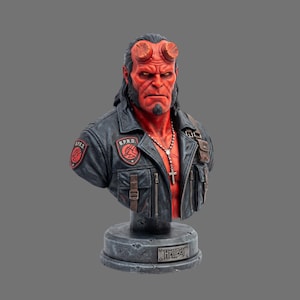Puede incluir: Un busto detallado del personaje Hellboy, con piel roja, cuernos prominentes y perilla. La figura viste una chaqueta negra con parches de B.P.R.D. en los hombros y el pecho, y un collar de cruz plateado. Se apoya sobre una base circular gris.