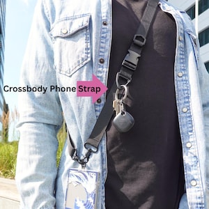 Op de afbeelding: Een zwarte crossbody telefoonriem met een zilveren clip en een zwarte sleutelhanger met een klein zwart apparaat. De riem wordt over de schouder van een persoon gedragen en is gelabeld "Crossbody Phone Strap".