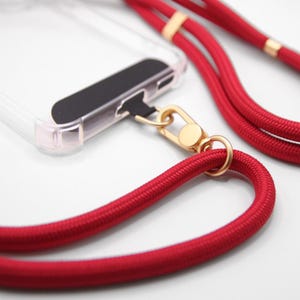 Puede incluir: Una correa de teléfono roja con herrajes dorados. La correa está hecha de un cordón grueso trenzado y tiene un broche dorado que se une a una funda de teléfono.