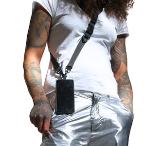 Könnte beinhalten: Eine Person trägt ein weißes T-Shirt und eine silberne Hose mit einer schwarzen Handyhülle, die an einem schwarzen Band befestigt ist, das über den Körper getragen wird.