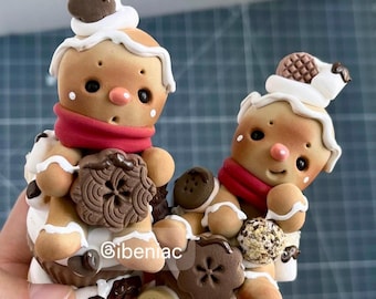 May include: Two tan gingerbread-style figures with red scarves, white icing details, and dark brown cookie accents. The figures are decorated with small round pink noses and black eyes. The text @ibeniac is visible on the front.