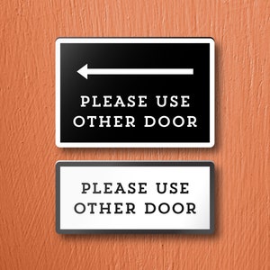 May include: Two black and white signs with white text on a black background. The top sign has a white arrow pointing to the left and the text "PLEASE USE OTHER DOOR". The bottom sign has the same text but no arrow.