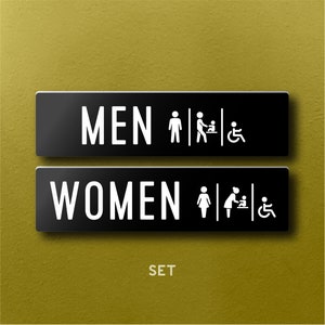 Op de afbeelding: Een set van twee zwarte rechthoekige toiletborden met witte tekst en pictogrammen. Het bovenste bord leest "MEN" met een stokfiguur, een pictogram voor het verschonen van baby's en een rolstoelsymbool. Het onderste bord leest "WOMEN" met vergelijkbare pictogrammen. Het woord "SET" staat eronder.