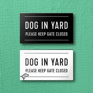 Op de afbeelding: Twee borden, een zwart en een wit, met de tekst "DOG IN YARD PLEASE KEEP GATE CLOSED". Het witte bord heeft een klein zwart silhouet van een hond in de linkerbenedenhoek.