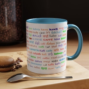 Könnte beinhalten: Weiße Keramik-Kaffeetasse mit blauem Innenraum und Henkel. Die Tasse ist mit dem Wort "Kaffee" in mehreren Sprachen bedruckt. Ein Silberlöffel und ein Macaron liegen auf einer Holzoberfläche.