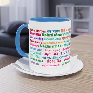 Op de afbeelding: Witte keramische mok met blauwe binnenkant en oor, met de tekst "Good Morning" in meerdere talen. De mok staat op een witte schotel, perfect voor koffie.