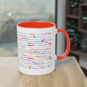 Könnte beinhalten: Eine weiße Keramiktasse mit rotem Innenraum und Griff. Die Tasse ist mit dem Wort "Hallo" in mehreren Sprachen in verschiedenen Farben verziert. Die Tasse ist für Heißgetränke konzipiert.