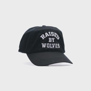 May include: Black baseball cap with the text "RAISED BY WOLVES" embroidered in white. The cap has a curved brim and a rounded crown. A casual accessory for everyday wear.