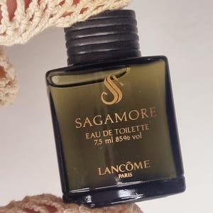 Puede incluir: Un pequeño frasco rectangular de perfume Sagamore Eau de Toilette. El frasco es de color verde oscuro con una tapa negra y letras doradas. El texto en el frasco dice "SAGAMORE EAU DE TOILETTE 7,5 ml 85% vol LANCÔME PARIS".