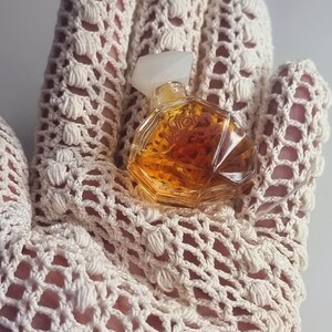 May include: A small, vintage perfume bottle with a clear, faceted design and a white cap. The bottle contains a golden-colored liquid. The bottle is resting on a cream-colored, open-weave crochet glove.