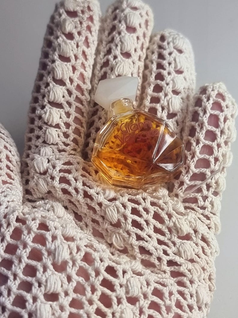 May include: A small, vintage perfume bottle with a clear, faceted design and a white cap. The bottle contains a golden-colored liquid. The bottle is resting on a cream-colored, open-weave crochet glove.