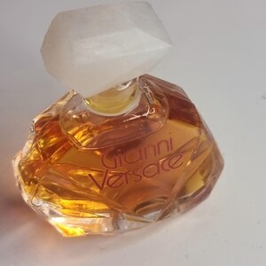 May include: A small, faceted perfume bottle with a clear, geometric stopper. The bottle contains a golden-colored liquid and features the name "Gianni Versace" in a reddish-brown font. The bottle has a unique, angular shape.