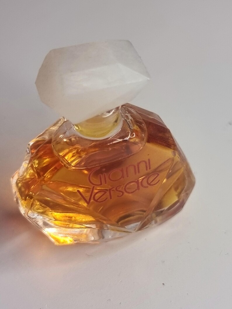 May include: A small, faceted perfume bottle with a clear, geometric stopper. The bottle contains a golden-colored liquid and features the name "Gianni Versace" in a reddish-brown font. The bottle has a unique, angular shape.