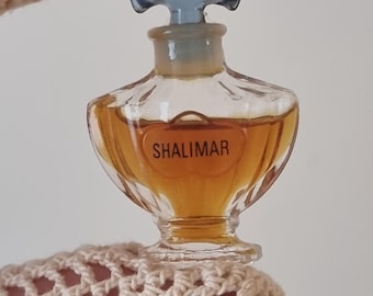 May include: A miniature perfume bottle of Shalimar by Guerlain. The clear glass bottle contains amber-colored liquid and features a light blue stopper. The label reads "SHALIMAR" in a classic font. The bottle is held by a crocheted sleeve.