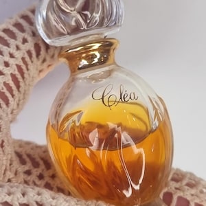 May include: A clear glass bottle of perfume with a gold cap and a gold label that says "Cléa". The bottle is filled with a yellow liquid.