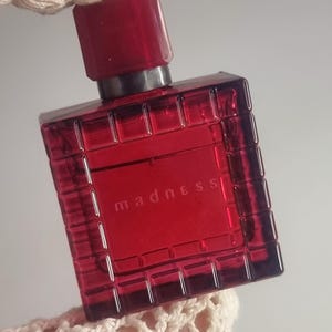 Könnte beinhalten: Eine rote Parfümflasche mit passendem Verschluss. Die rechteckige Flasche hat ein gitterartiges Muster und das Wort "madness" ist auf der Vorderseite aufgedruckt. Die Flasche wird von einer cremefarbenen Häkelmanschette gehalten.