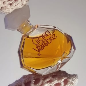May include: A small, faceted perfume bottle with a clear, geometric design. The bottle contains a golden-colored liquid and features the text "Gianni Versace" in red lettering. The bottle has a white cap and is held by a crocheted fabric.