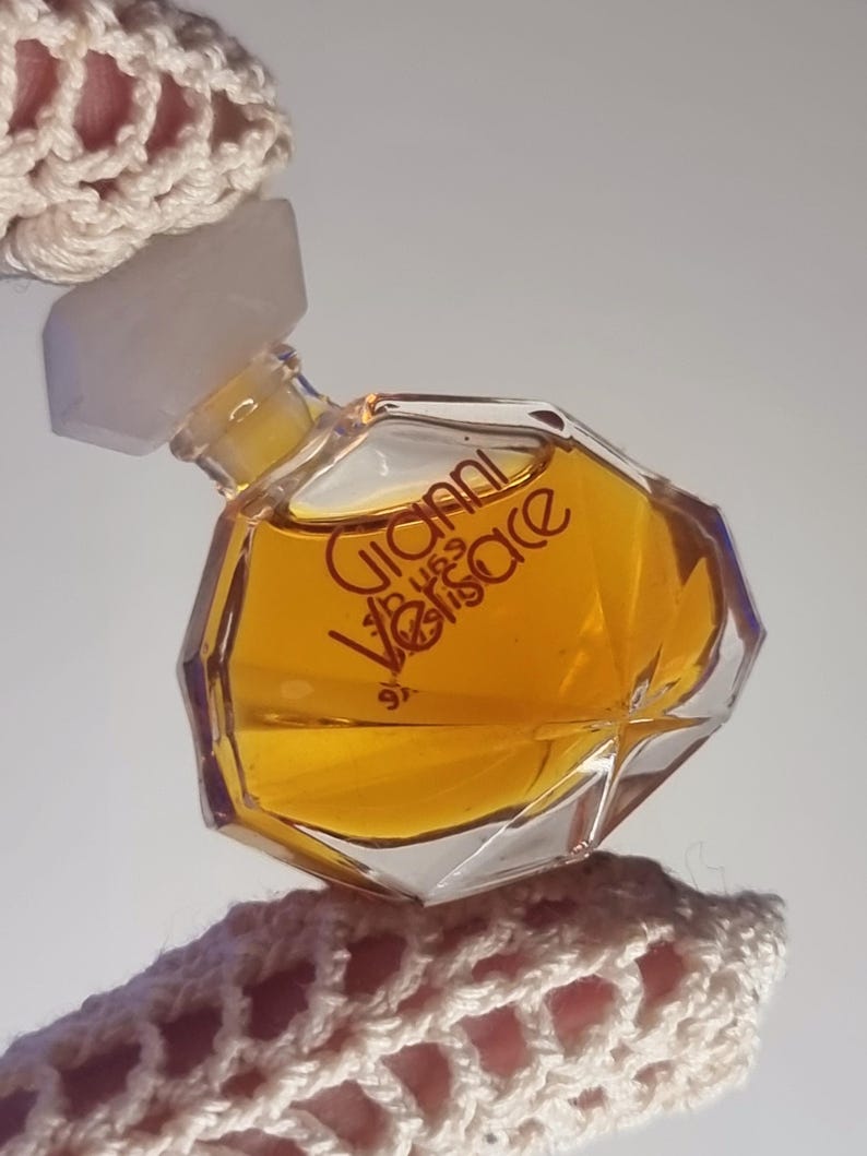 May include: A small, faceted perfume bottle with a clear, geometric design. The bottle contains a golden-colored liquid and features the text "Gianni Versace" in red lettering. The bottle has a white cap and is held by a crocheted fabric.