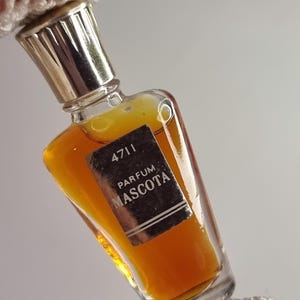 Pode incluir: Um pequeno frasco de perfume vintage com uma tampa dourada e um rótulo retangular. O rótulo diz "4711 PARFUM MASCOTA" em texto preto. O frasco contém um líquido amarelo dourado.