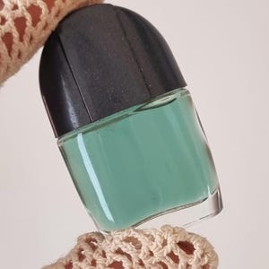 Könnte beinhalten: Eine klare Glas-Nagellackflasche, gefüllt mit einem helltürkisfarbenen Lack. Die Flasche hat einen schwarzen Deckel. Die Flasche wird von einem gehäkelten Ärmel gehalten.
