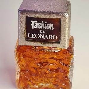 Op de afbeelding: Een vintage parfumflesje met een zilverkleurige dop en een getextureerde, amberkleurige glazen body. Het etiket leest "Fashion DE LEONARD" in witte letters op een bruine achtergrond. De fles heeft een uniek, artistiek ontwerp.