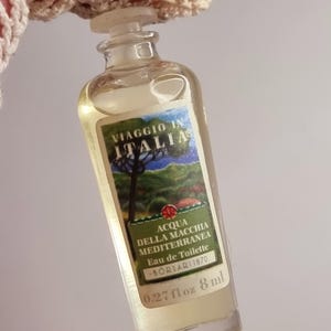 May include: A small, clear glass perfume bottle with a label featuring the text "VIAGGIO IN ITALIA" and "ACQUA DELLA MACCHIA MEDITERRANEA". The bottle contains a light yellow liquid and is 0.27 fl oz (8 ml).