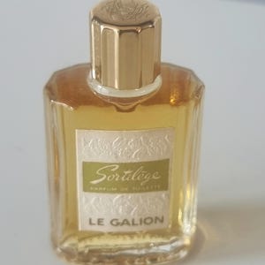 May include: A small, clear glass bottle of perfume with a gold cap. The bottle has a label that reads "Sortilege Parfum de Toilette Le Galion".