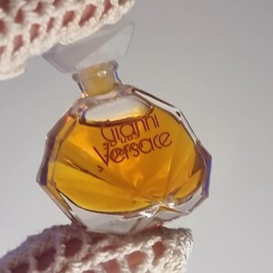 May include: A small, octagonal perfume bottle filled with amber liquid. The bottle has a clear plastic cap and the words "Gianni Versace" are printed in gold. The bottle is held by a crocheted fabric.