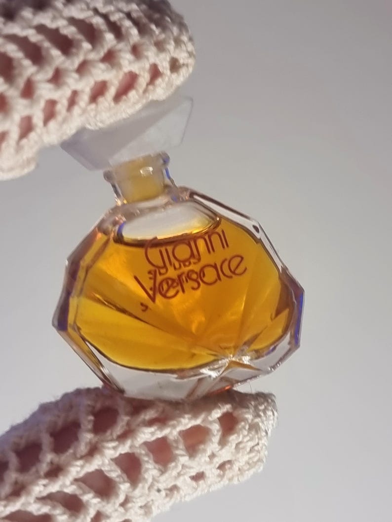 May include: A small, octagonal perfume bottle filled with amber liquid. The bottle has a clear plastic cap and the words "Gianni Versace" are printed in gold. The bottle is held by a crocheted fabric.