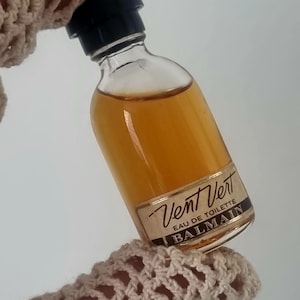 Puede incluir: Un pequeño frasco de vidrio de Vent Vert Eau de Toilette de Balmain. El frasco está lleno de un líquido dorado y tiene una tapa negra. La etiqueta es blanca con letras negras.