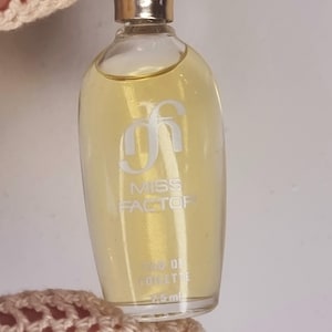 Op de afbeelding: Een klein, doorzichtig glazen flesje Miss Factor Eau de Toilette. Het flesje is gevuld met een gele vloeistof en heeft een gouden dop. Het flesje is gelabeld met de woorden "Miss Factor" en "Eau de Toilette".