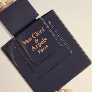 May include: A dark navy blue rectangular perfume bottle with a matching square cap. The front of the bottle features the gold-colored text Van Cleef & Arpels Paris embossed in the center. The bottle has a smooth, glossy finish and a clean design.