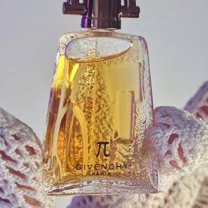 May include: A clear glass perfume bottle with a trapezoidal shape and a dark metallic cap. The bottle contains a golden liquid and features the Greek letter Pi symbol above the text GIVENCHY PARIS printed in black on the front surface.
