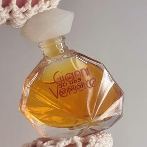 May include: A small, clear glass perfume bottle with a geometric shape, filled with a golden-colored liquid. The bottle has a white, faceted cap and the name "Gianni Versace" is printed on the front. The bottle is held by a hand wearing a cream-colored crocheted sleeve.