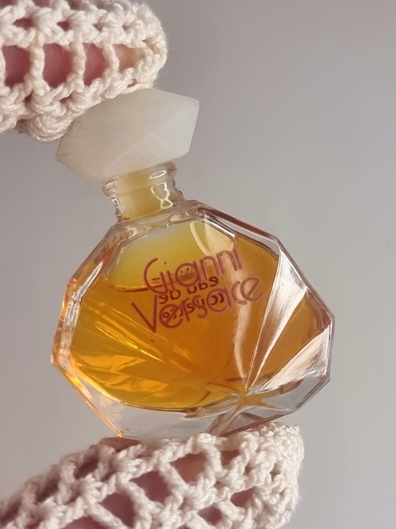 May include: A small, clear glass perfume bottle with a geometric shape, filled with a golden-colored liquid. The bottle has a white, faceted cap and the name "Gianni Versace" is printed on the front. The bottle is held by a hand wearing a cream-colored crocheted sleeve.