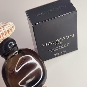 May include: A dark brown perfume bottle and a black box with the name "HALSTON" printed on them. The box also has the text "HALSTON 1-12 EAU DE TOILETTE POUR HOMME 12 ml". The bottle is round and sits on a cream-colored crocheted surface.