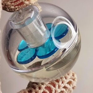 May include: A clear, spherical perfume bottle with a transparent plastic top. Inside, there are several blue, circular accents. The word "Pupa" is visible on the blue accents. The bottle is held by a beige crocheted item.
