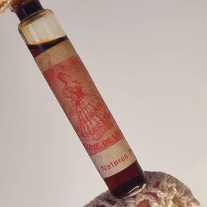 May include: A vintage perfume bottle with a red and cream label featuring an illustration of a woman in a dress. The bottle is filled with a dark amber liquid. The label reads "Parfume de Las Natures P".