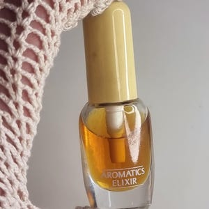 Może przedstawiać: Mała, przezroczysta szklana butelka perfum z żółtą nakrętką. Butelka zawiera płyn w kolorze złotym i ma napis "AROMATICS ELIXIR" z przodu. Butelka jest trzymana przez kremowy, szydełkowy rękaw.