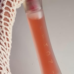 May include: A frosted glass perfume bottle with a pink liquid inside. The bottle has a clear cap and gold lettering that reads "ZOLTA". The bottle is held by a cream-colored crocheted sleeve.
