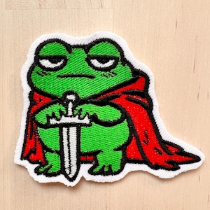 Peut inclure: Écusson brodé représentant une grenouille verte portant une cape rouge et tenant une épée argentée. La grenouille a une expression sévère et est bordée de noir. L'écusson a une bordure blanche.