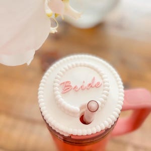 May include: A pink tumbler with a white lid featuring the word "Bride" in pink script. The lid has a beaded edge and a straw. A white orchid flower is in the background.