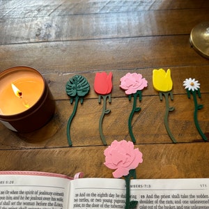 May include: A set of six colorful flower-shaped bookmarks. The bookmarks are made of plastic and feature a variety of flowers, including a tulip, a daisy, a carnation, and a monstera leaf. The bookmarks are all green on the back and have a long stem.