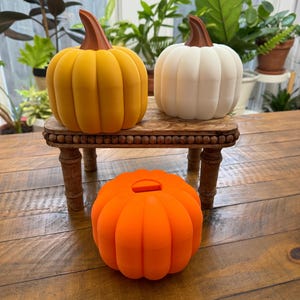 以下が含まれることがあります： 黄色、白、オレンジの3つの装飾的なセラミック製かぼちゃが、木製スタンドに飾られています。