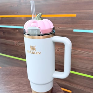 以下が含まれることがあります： 金色のベースとハンドルが付いた白いスタンレータンブラー。ピンクのかぼちゃ型の蓋と透明なストローが付いています。スタンレーのロゴは金色です。タンブラーは木製の表面に置かれています。
