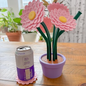 Puede incluir: Una maceta rosa con tres flores rosas y tallos verdes. La maceta es morada y tiene una base marrón. Una lata de bebida con sabor a uva está sobre un posavasos en forma de flor rosa.