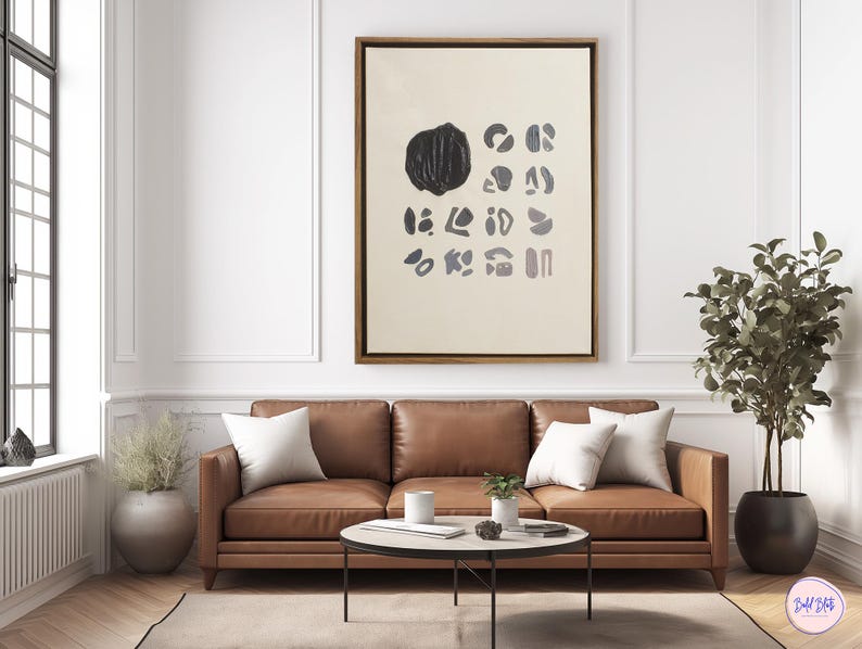 May include: A brown leather sofa with two white pillows in front of a beige wall with a framed abstract painting featuring black and gray shapes. A potted plant with green leaves is on the right side of the image.